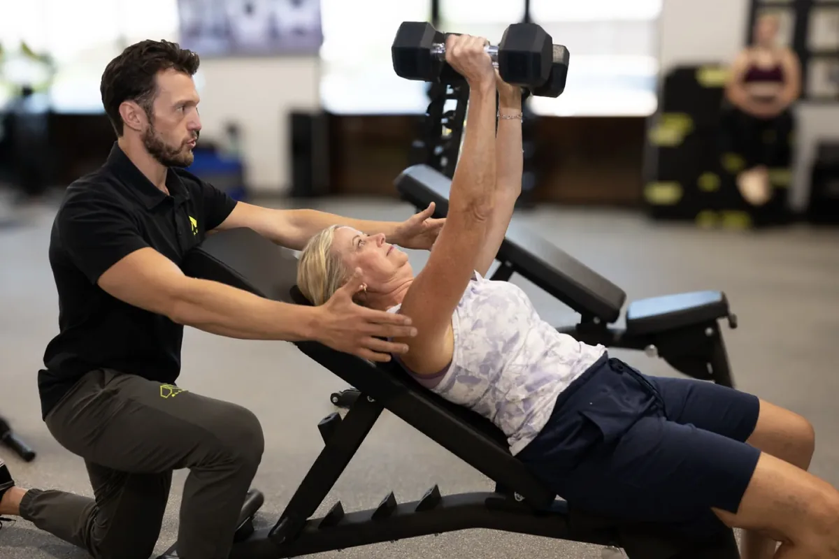 A trainer guides a member through an incline dumbbell press during a coached session